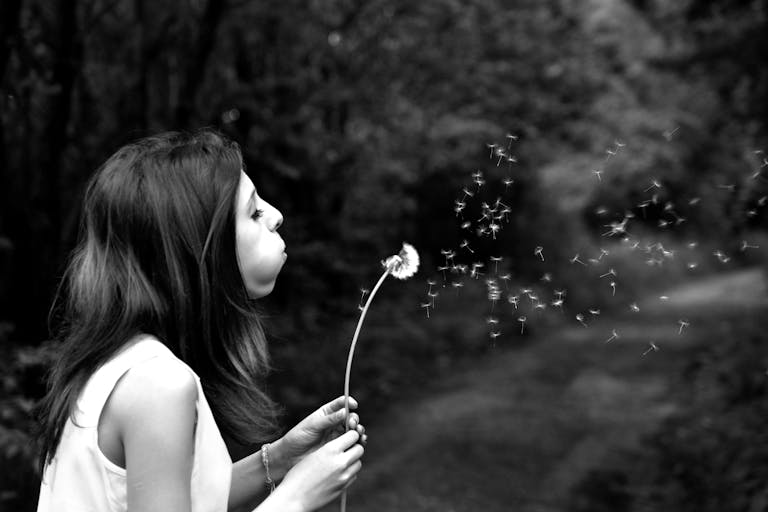 A woman blows dandelion seeds in a serene forest, captured in black and white.
